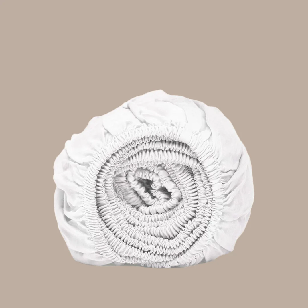 Opgerold wit hoeslaken van bamboe, zachte stof. Neutrale achtergrond, zacht licht. Elastiek detail zichtbaar.