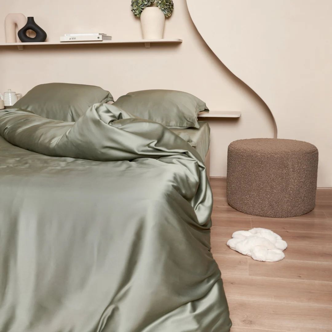 Olijfgroen bamboe hoeslaken opgemaakt op een bed, gezien vanuit een laag standpunt. Zachte, natuurlijke belichting en warme achtergrondkleuren.