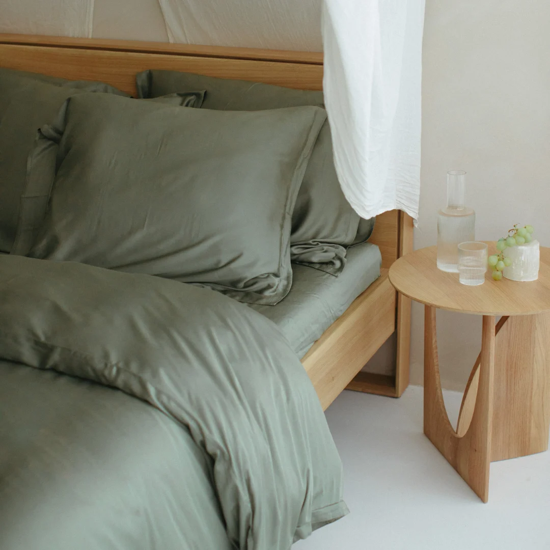 Olijfgroen bamboe hoeslaken op houten bedframe, zacht licht. Kussen en dekbedovertrek tonen de textuur. Houten nachtkastje met water en druiven.