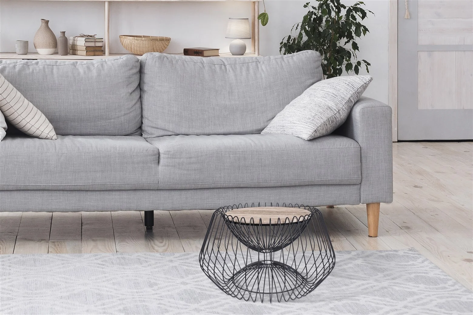 Een moderne woonkamer is voorzien van een lichtgrijze bank, twee gedessineerde kussens, een Bijzettafel Zwart, Bruin 37 CM Rond marmer salontafel, een lichtgrijs vloerkleed, een plant in pot en decorplanken. De ruimte heeft een lichte, minimale stijl.