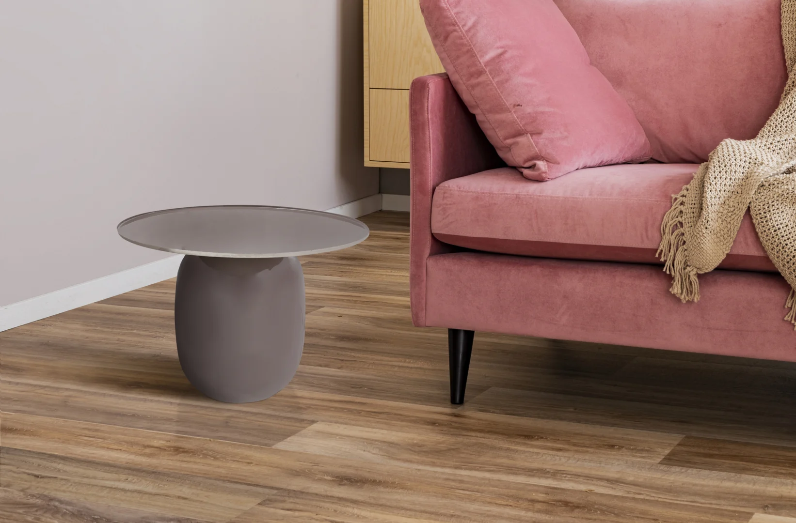 Een moderne woonkamer met een roze fluwelen bank, een beige gebreide plaid, een Bijzettafel Zilver (klein grijs rond ijzeren bijzettafeltje), een houten vloer en een licht houten kast op de achtergrond.