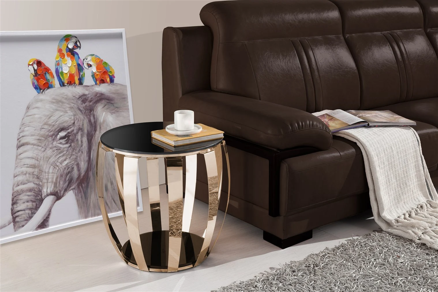 Een moderne woonkamer toont een bruine leren bank met een beige kleed en open boek, een Bijzettafel Goud Staal Compact met een kaars en boek, een grijs vloerkleed en een ingelijst olifantenkunstwerk tegen de muur.