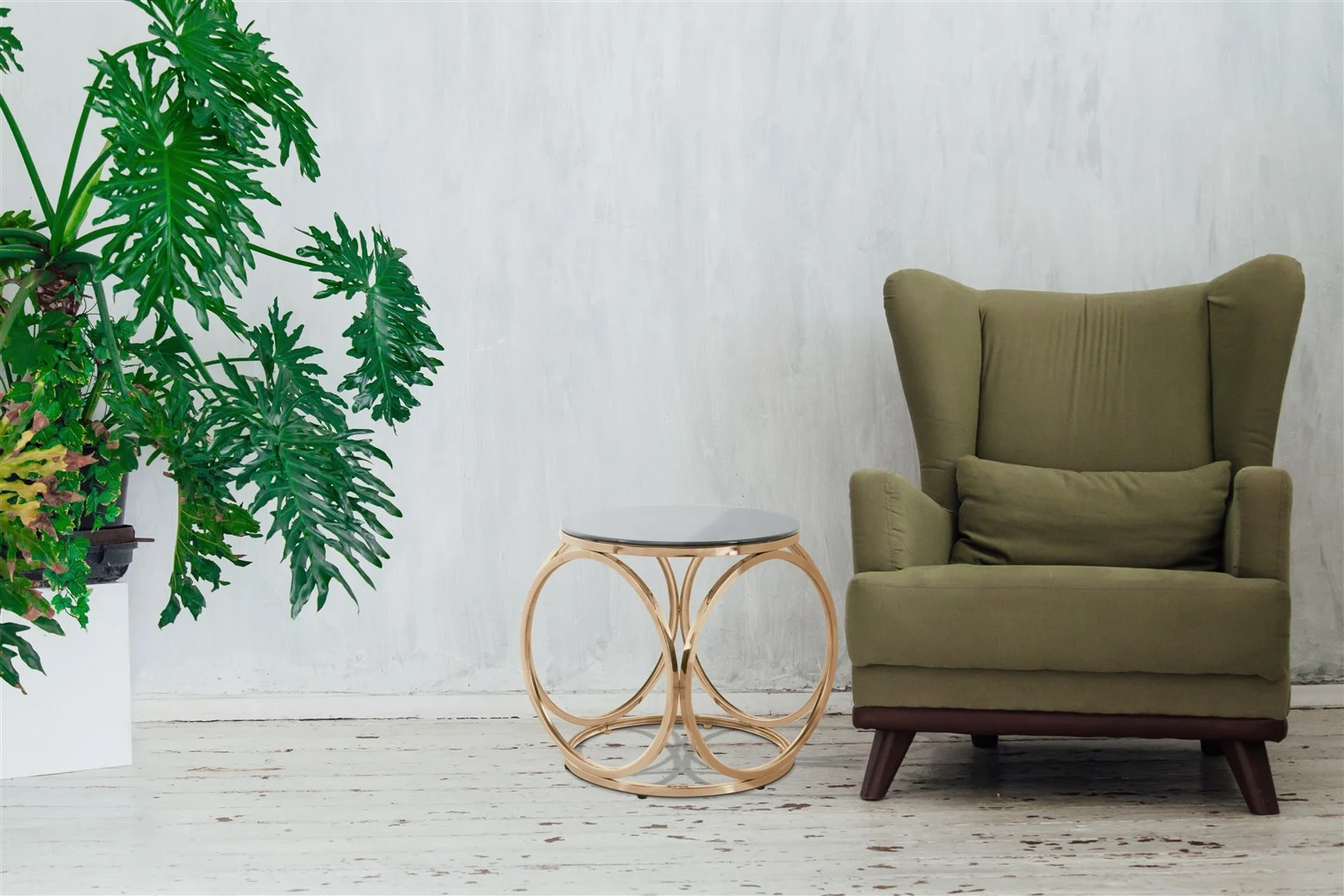 Een groene fauteuil met kussen staat naast de Bijzettafel Grijs Staal Compact en een grote bladplant in een witte pot, tegen een lichtgrijze muur met structuur en een witte houten vloer.