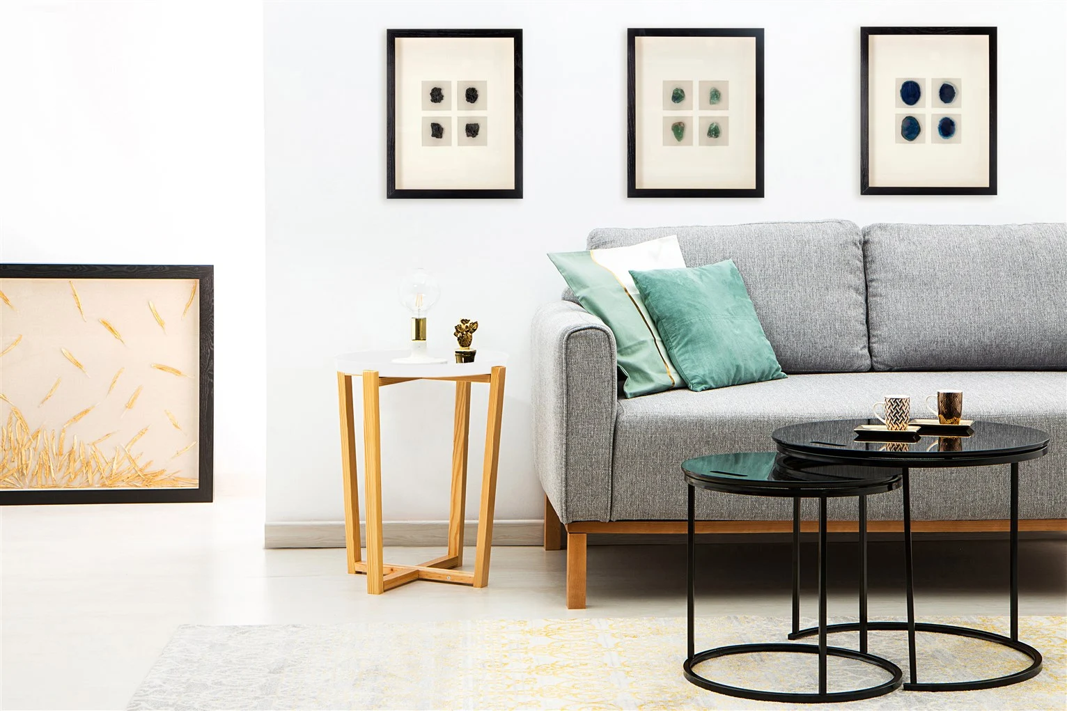 Een moderne woonkamer met een grijze bank, groene en witte kussens, twee ronde zwarte salontafels, de Bijzettafel Wit Grenenhout Design met decor, ingelijste kunst aan de muur en muurkunst op de vloer.