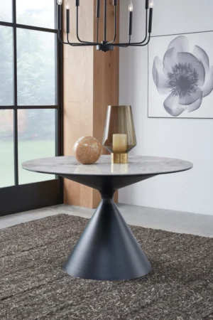 De Winston stenen blad metalen onderstel ronde eettafel in Grigio, met een marmeren blad en zwart metalen onderstel, staat op een ruig tapijt met een glazen vaas, bol en kaars. Een groot bloemenkunstwerk en een raam maken het tafereel compleet.