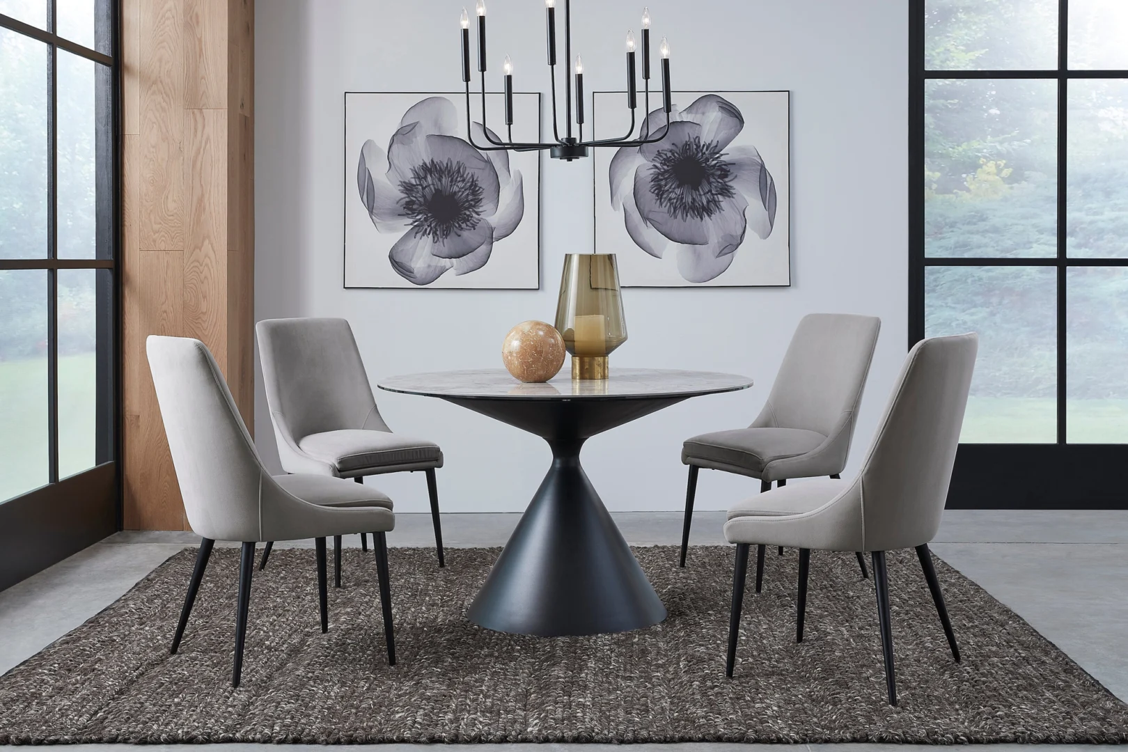 Een moderne eetkamer met de Winston stenen blad metalen onderstel ronde eettafel in Grigio, gecombineerd met vier lichtgrijze stoelen, een vloerkleed met donkere structuur, een zwarte kroonluchter, decoratieve vazen, bloemenkunst en grote ramen die natuurlijk licht binnenlaten.