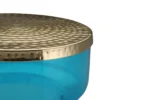 Een close-up van de Bijzettafel Blauw, met een gestructureerd gouden metalen deksel en een doorschijnend blauw rond lichaam, tegen een witte achtergrond.