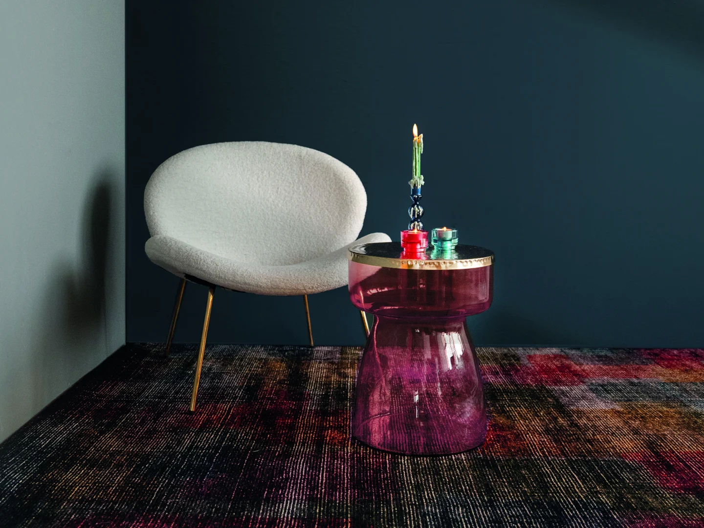 Een moderne witte stoel met gouden poten staat naast de Bijzettafel Goud, Roze Ijzer Compact, met daarop twee brandende kaarsen, allemaal gerangschikt op een gestructureerd, veelkleurig tapijt tegen een donkerblauwe muur.