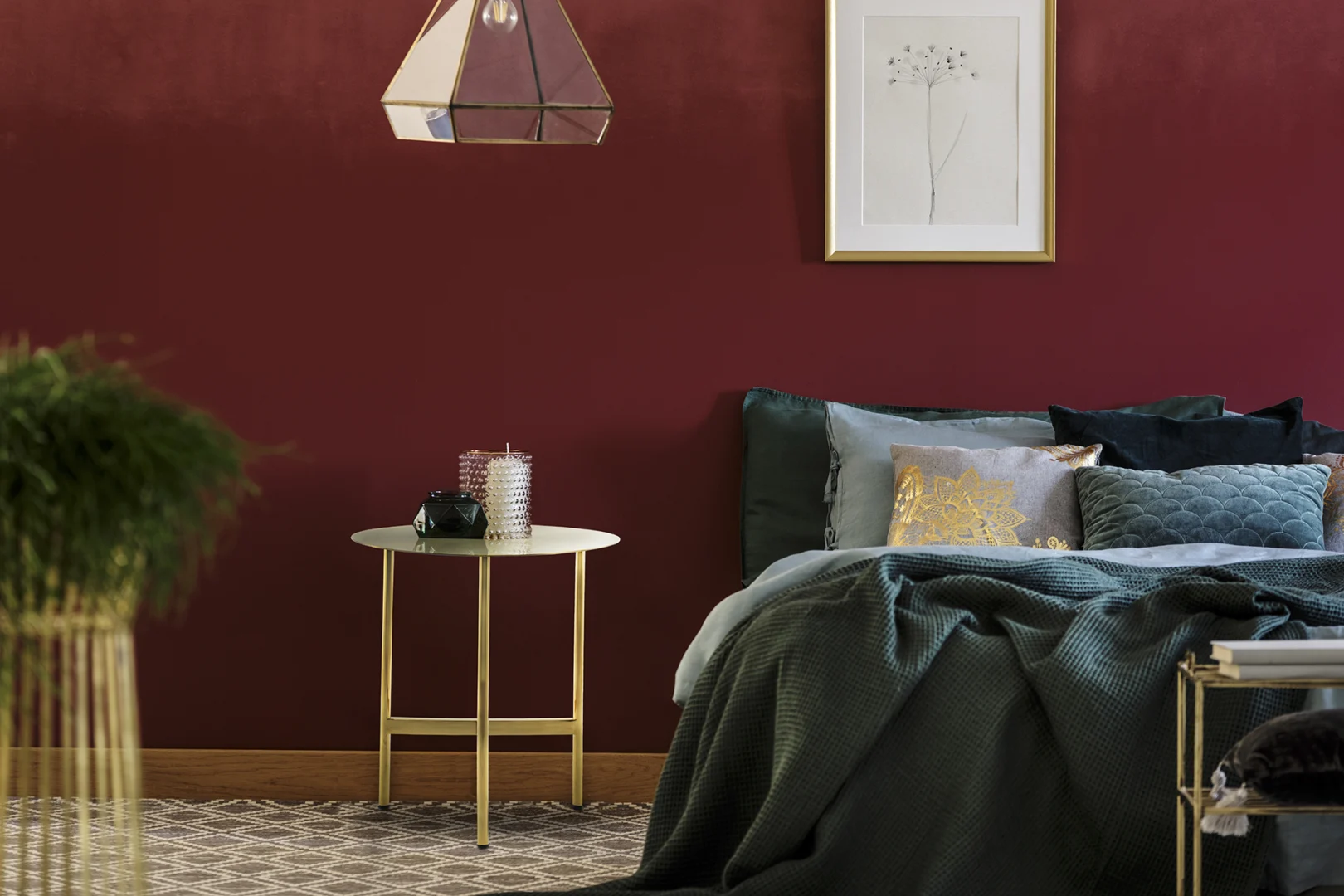 Een gezellige slaapkamer met een donkerrode muur, een ingelijste botanische print, een bed met groenblauwe en gouden kussens, een groene deken en de Bijzettafel Goud Rond Staal Compact naast het bed met decoratiespullen.