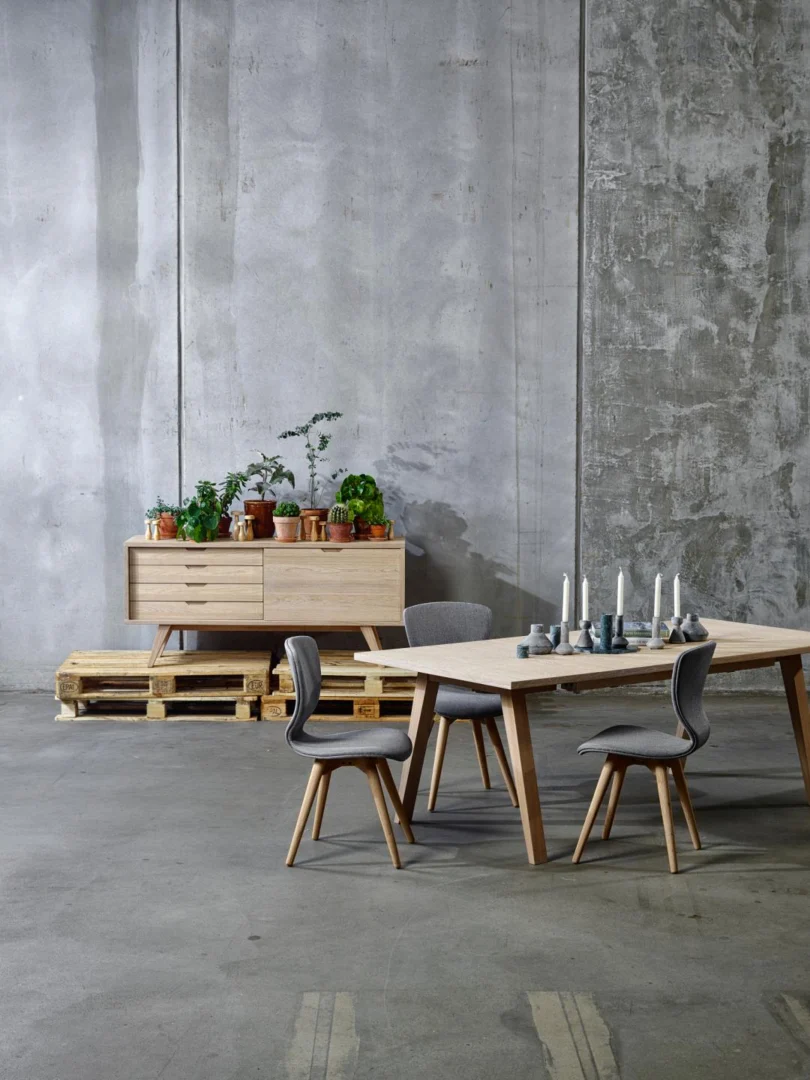 Een minimalistische eethoek met de Eettafel Nieuwe Sfeer 100 x 74 CM, een rechthoekige eiken tafel, gekoppeld aan vier grijze stoelen en een modern dressoir met planten in potten, alles tegen kale betonnen muren en vloeren voor een industriële look.