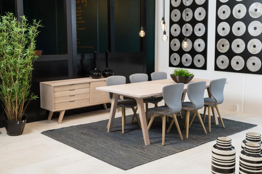 Een moderne eetkamer met de Eettafel Nieuwe Sfeer 100 x 74 CM rechthoek eikenhout tafel, zes grijs gestoffeerde stoelen, een donker vloerkleed, een dressoir, hanglampen, abstracte wandkunst, bamboe in potten en gestreepte decoratieve vazen.