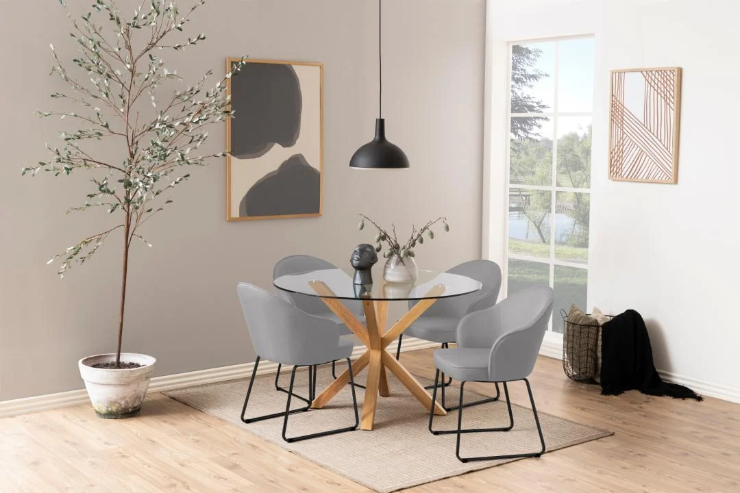 In een moderne eetkamer staat de Eettafel Nieuwe Sfeer 119 CM Rond Glas met een stalen onderstel, vier grijze stoelen, een potplant, abstracte muurkunst, een zwarte hanglamp en een raam voor natuurlijk licht. Neutrale tinten en een minimalistische inrichting maken de look compleet.