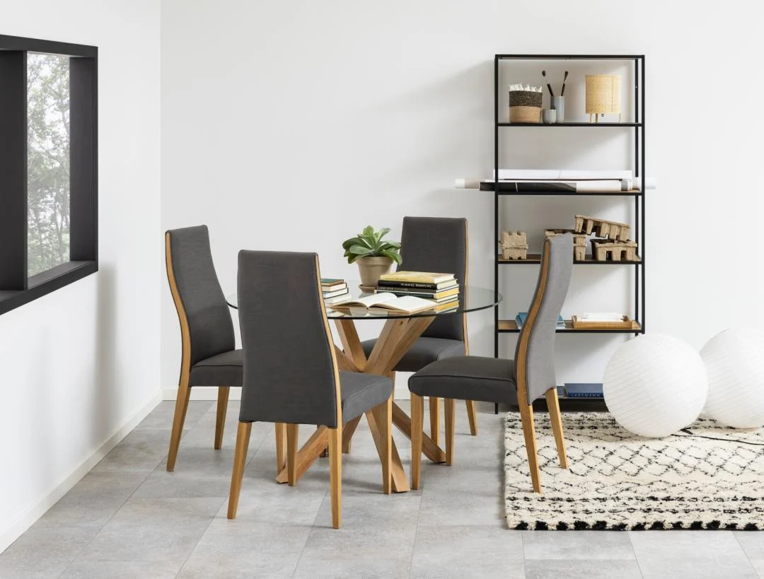 Een moderne eethoek met de Eettafel Nieuwe Sfeer 119 CM Rond Glas met een stalen onderstel, vier grijze stoelen, een zwarte metalen plank met decor, twee witte staande lampen, een vloerkleed met patroon op een tegelvloer, plus een plant en boeken op tafel.