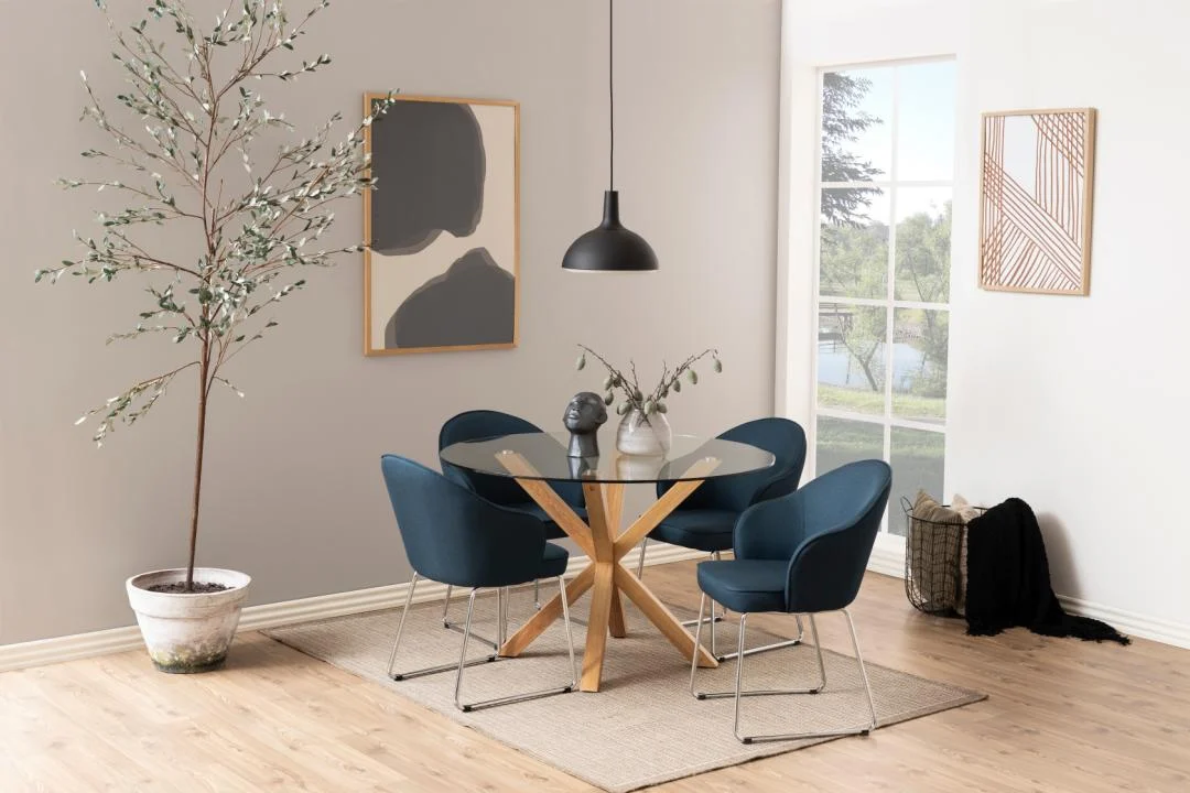 Een moderne eetkamer met de Eettafel Nieuwe Sfeer 119 CM Rond Glas met stalen frame, vier donkerblauwe stoelen, een zwarte hanglamp, abstracte muurkunst, een hoge potplant en een groot raam dat natuurlijk licht binnenlaat.
