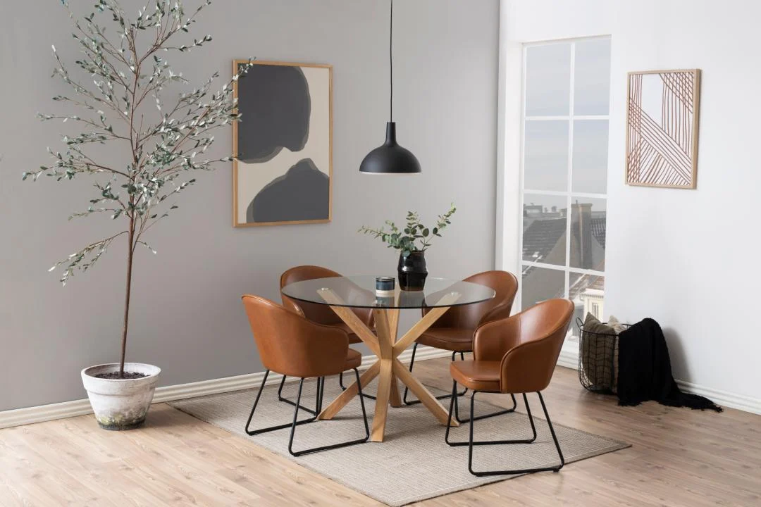 Moderne eetkamer met de Eettafel Nieuwe Sfeer 119 CM Rond Glas met een stalen onderstel, vier bruine leren stoelen, een grote potplant, abstracte muurkunst, een zwarte hanglamp en een raam met uitzicht op de stad.