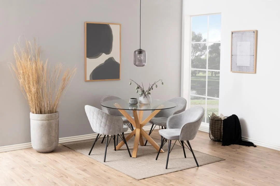Een moderne eethoek met de Eettafel Nieuwe Sfeer 119 CM Rond Glas met een stalen onderstel, vier grijze stoelen met kussens, een hanglamp, abstracte muurkunst, een hoge vaas met gedroogd gras, een raam en een geweven dekenmand op een lichthouten vloer.