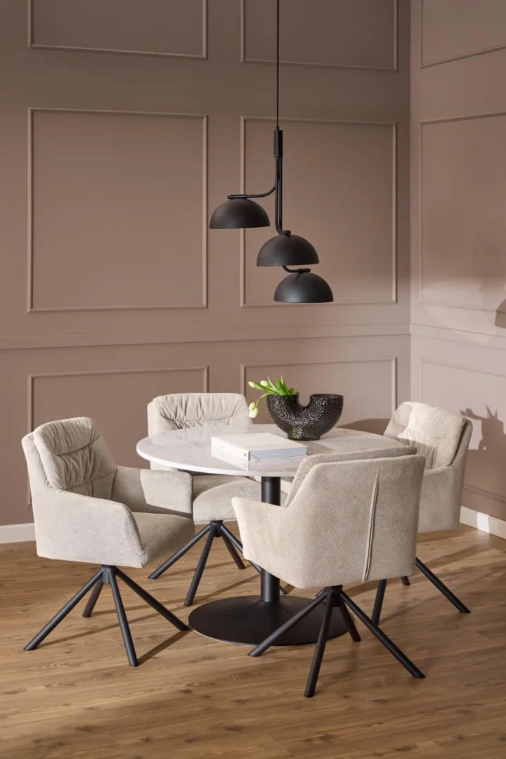 Een moderne eethoek met de Eettafel Nieuwe Sfeer 110 CM Rond Marmer, een matzwarte basis, vier beige stoelen met kussens, een zwarte schaal en boek op tafel, een houten vloer, muren met neutrale panelen en drie zwarte hanglampen erboven.