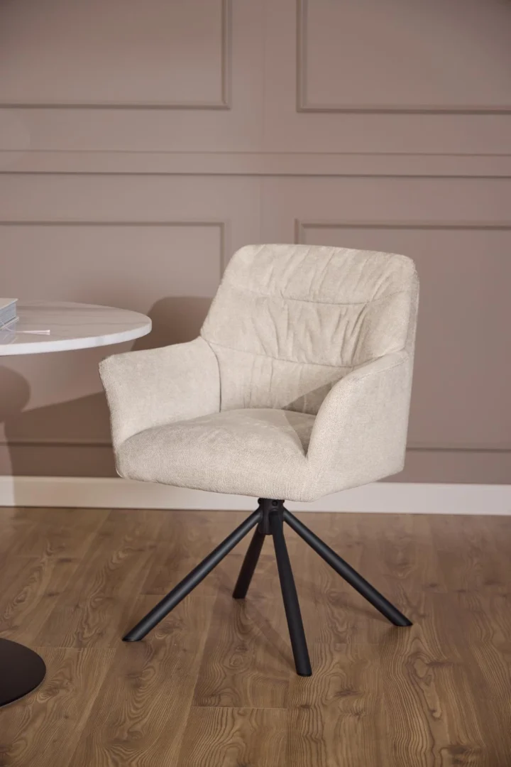 Een moderne beige gestoffeerde stoel met armleuningen staat op een houten vloer naast de Eettafel Nieuwe Sfeer 110 CM Rond Marmer, die is voorzien van een rond marmeren blad en een mat zwart onderstel, tegen een wand met taupe panelen.