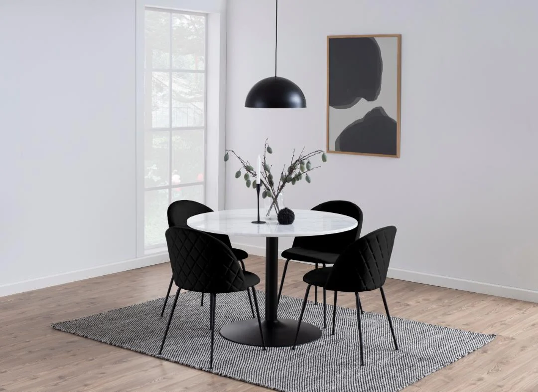 Een moderne eethoek met de Eettafel Nieuwe Sfeer 110 CM Rond Marmer met een matzwart onderstel, vier zwart gewatteerde stoelen, een zwarte hanglamp, een vaas met takken, abstracte muurkunst en een groot raam op een tapijt met grijze structuur.