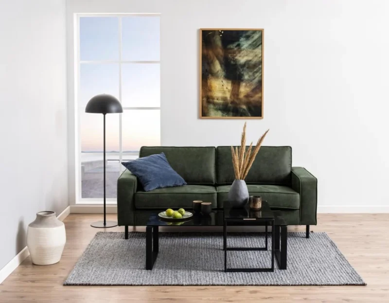 Een moderne woonkamer met een groene bank, zwarte staande lamp, abstracte muurkunst, blauw kussen, vaas met gedroogd gras, schaal met appels en een Salontafel Rookglas (zwart, 40 x 45 cm) staat bij een groot raam met uitzicht naar buiten.
