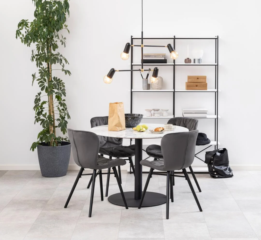 In een moderne eethoek staat de Eettafel Nieuwe Sfeer 105 CM Rond Marmer met een matzwart onderstel, vier grijze stoelen, een papieren zak met fruit en een zwarte schaal. Daarachter staat een zwart rek met boeken en decor, plus een hoge potplant aan de linkerkant.