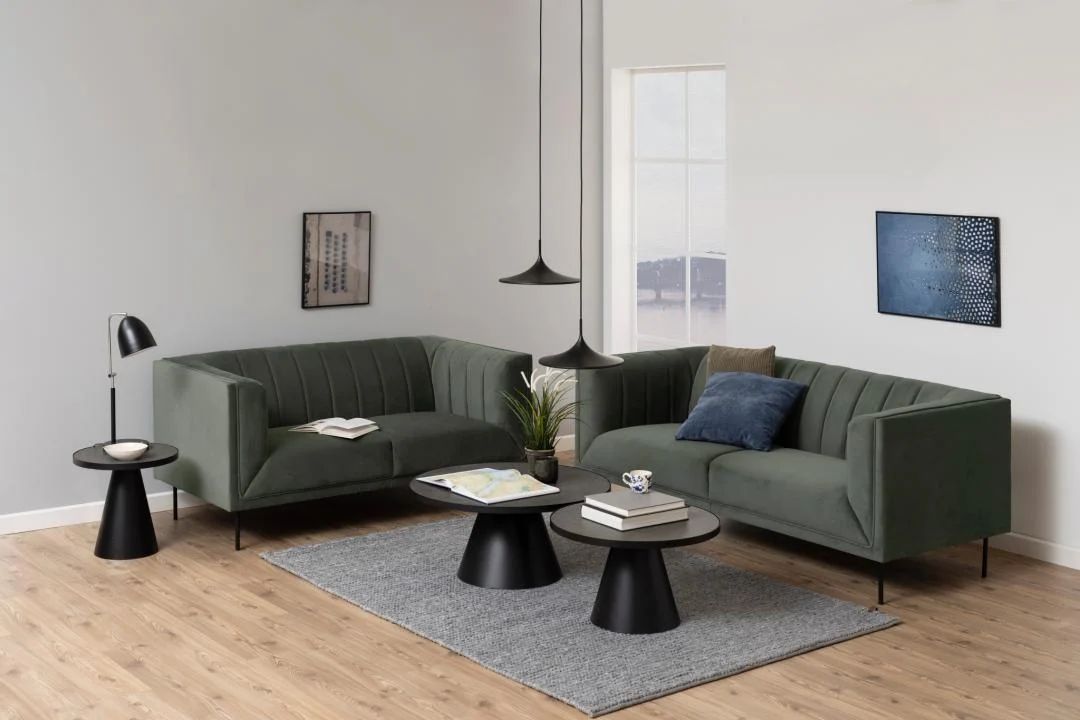 Een moderne woonkamer met twee groene banken, Salontafel Zwart 45 CM Rond Keramiek, een grijs vloerkleed, staande lamp, boeken, een plant en muurkunst. Licht van een hoog raam accentueert de minimalistische inrichting.