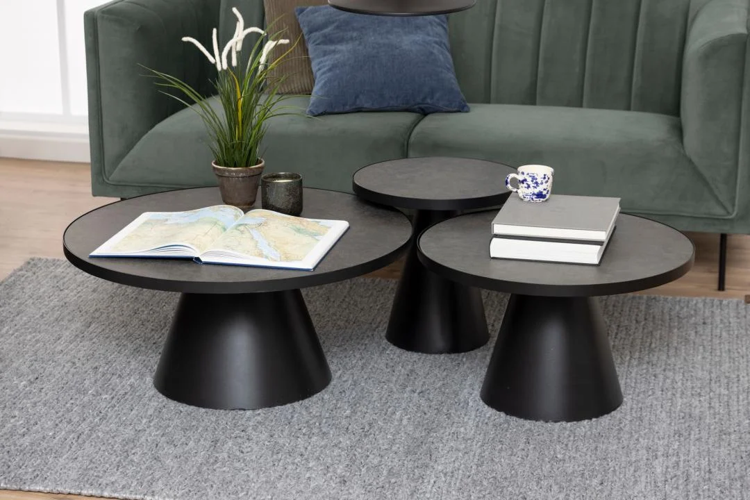 De Salontafel Zwart 45 CM Rond Keramiek toont een stapel boeken, een blauw-wit theekopje, een opengeslagen kaartenboek, twee kleine potplanten en een kaars op een grijs vloerkleed, met een groene bank op de achtergrond.