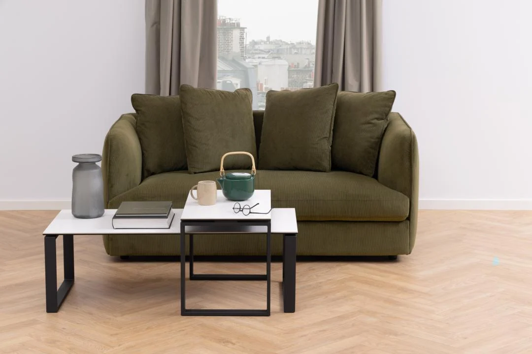 Een groene bank met vier kussens staat onder een raam met beige gordijnen. Aan de voorkant staan twee ronde keramische Salontafels Zwart, Wit 40 x 45 CM met een theepot, beker, glazen vaas en boeken op een lichthouten visgraatvloer.
