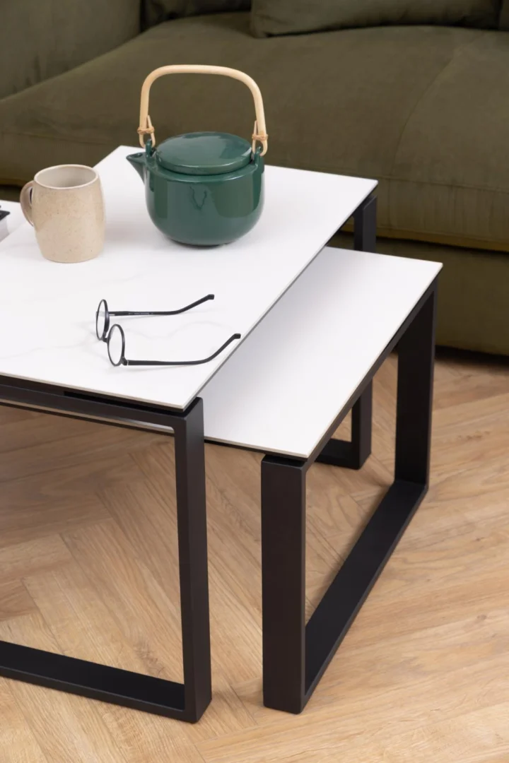 De Salontafel Zwart, Wit 40 x 45 CM Rond van keramiek staat voor een groene bank op een houten vloer en draagt een groene theepot, een beige mok en een bril.