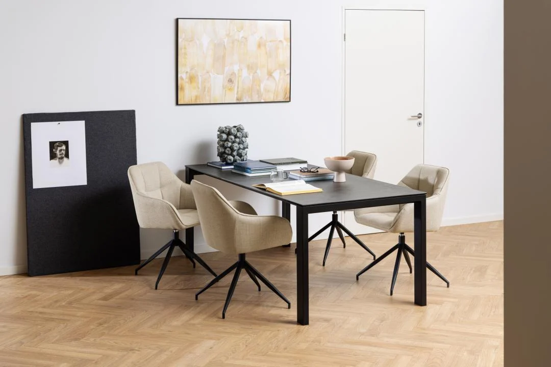 Een modern kantoor met de Eettafel Brentford 200 x 90 CM Rechthoek in zwart keramiek, vier beige bureaustoelen, boeken en decor op tafel. Kunstwerken sieren witte muren boven een lichthouten visgraatvloer.