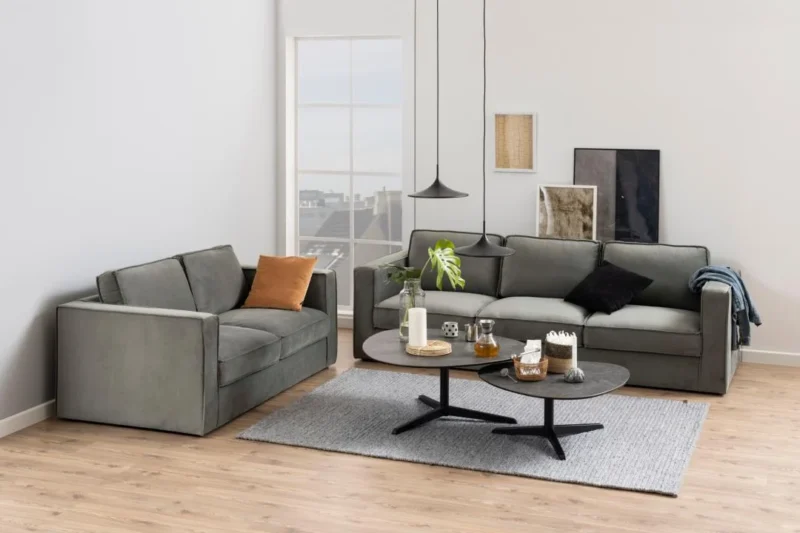 Een moderne woonkamer met twee grijze banken, een grijs vloerkleed en de Salontafel Zwart 95 x 41 CM Driehoek (kunststof) als salontafel met drankjes en decor. Neutrale muurkunst, een lichte houten vloer en een groot raam fleuren de ruimte op.