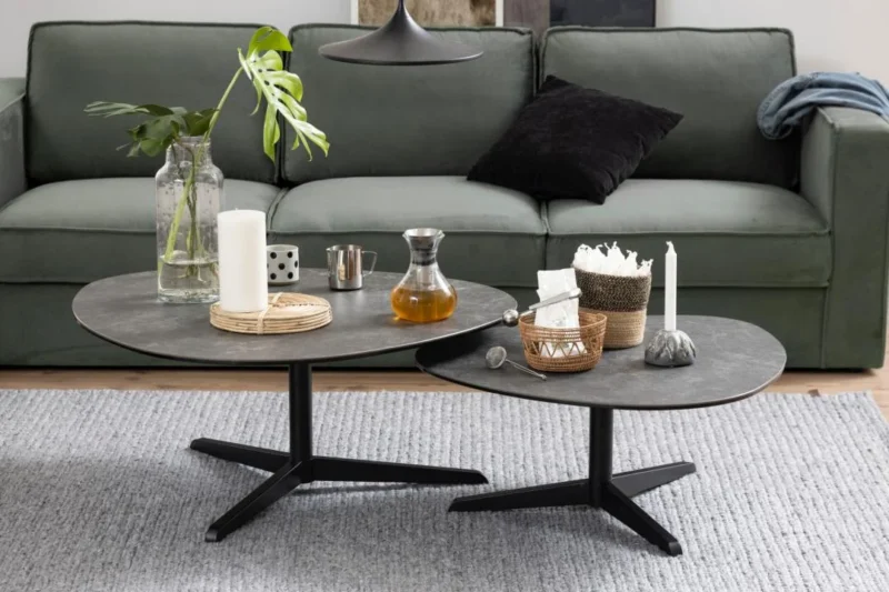 De Salontafel Zwart 95 x 41 CM Driehoek, gemaakt van kunststof, is gestyled met kaarsen, een glazen pot, een vaas met groene bladeren en een karaf, en geplaatst op een grijs vloerkleed voor een groene bank met zwarte en blauwe kussens.
