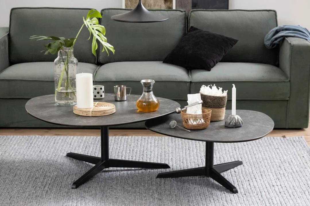 De Salontafel Zwart 95 x 41 CM Driehoek, gemaakt van kunststof, is gestyled met kaarsen, een glazen pot, een vaas met groene bladeren en een karaf, en geplaatst op een grijs vloerkleed voor een groene bank met zwarte en blauwe kussens.