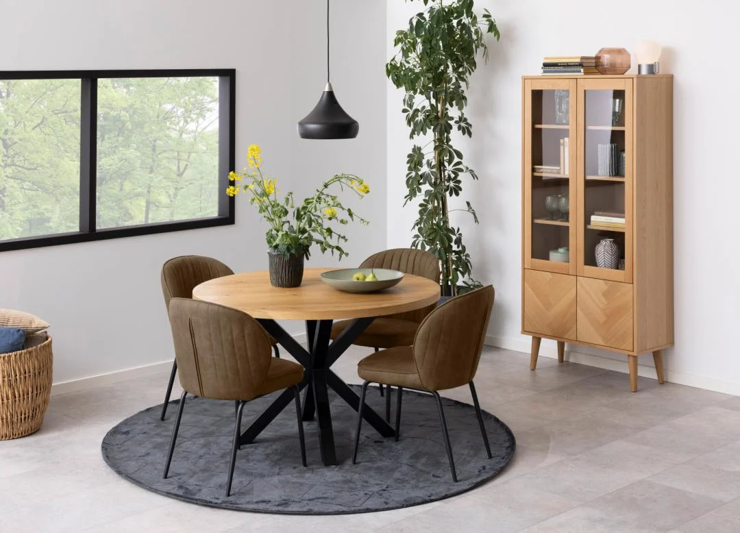 In een moderne eetkamer staan de Eettafel Hemel 120 CM Rond Eikenhout Staal, vier bruine stoelen, een grote groene plant, een zwarte hanglamp, een houten vitrinekast, groen voor het raam en een grijs vloerkleed onder de tafel.
