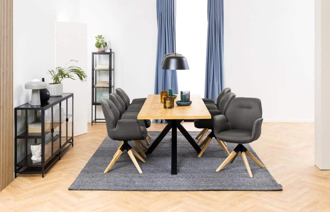 Een moderne eetkamer met de Eettafel Hemel 220 x 90 CM Rechthoek in eikenhout staal, acht grijze stoelen, een zwarte hanglamp, blauwe gordijnen, een grijs vloerkleed en planken met planten tegen witte muren en lichthouten vloeren.