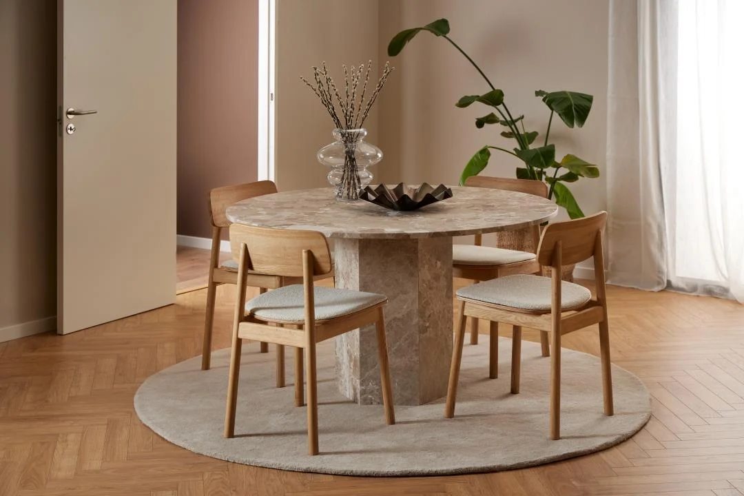 De Eettafel Naxos 130 CM Rond Marmer Massief is gecombineerd met vier houten stoelen op een rond vloerkleed in een moderne minimalistische eetkamer, met een vaas met gedroogde takken, een decoratieve schaal en een groene plant op de achtergrond.