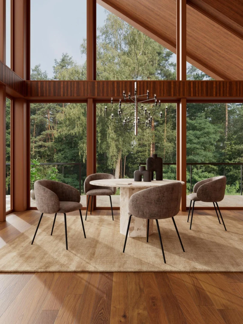Een moderne eethoek met de Eettafel Naxos 130 CM Rond Marmer Massief met vier grijze stoelen op een beige vloerkleed, bij ramen van vloer tot plafond met uitzicht op een bos en verlicht door een zwarte kroonluchter.