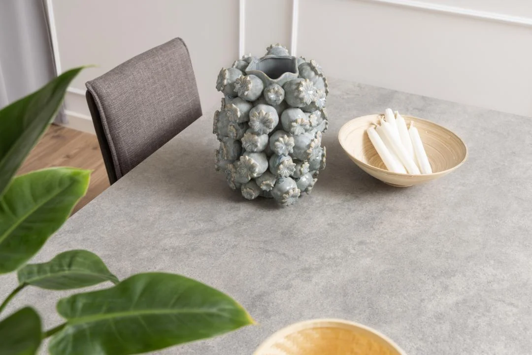 De Eettafel Nieuwe Sfeer 200 x 100 CM, een grijze rechthoekige tafel van keramiek en staal, is vormgegeven met een stekelige blauwgrijze vaas, beige schalen - één met witte kaarsen - een grijze stoel en een groene bladplant op de voorgrond.