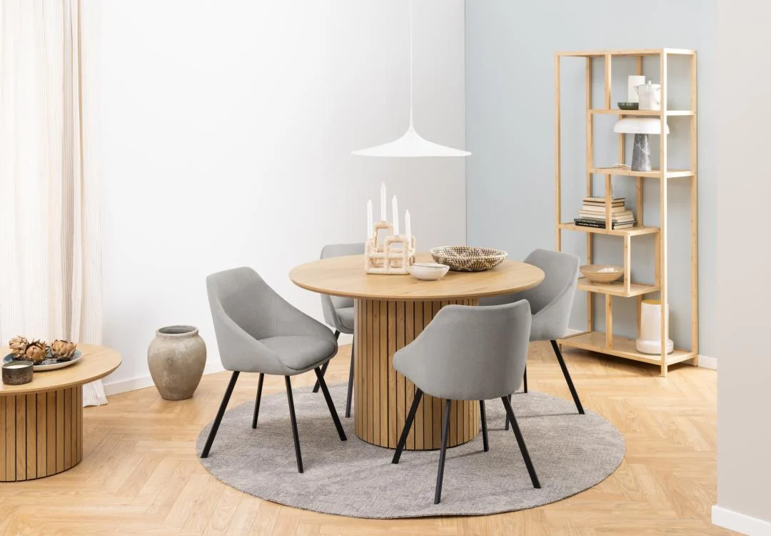 Een moderne eethoek met de Eettafel Nieuwe Sfeer 120 CM Rond MDF tafel, vier grijs gestoffeerde stoelen, een cirkelvormig vloerkleed, een houten stellingkast met decor en neutrale muren. Natuurlijk licht valt binnen vanaf de linkerkant.