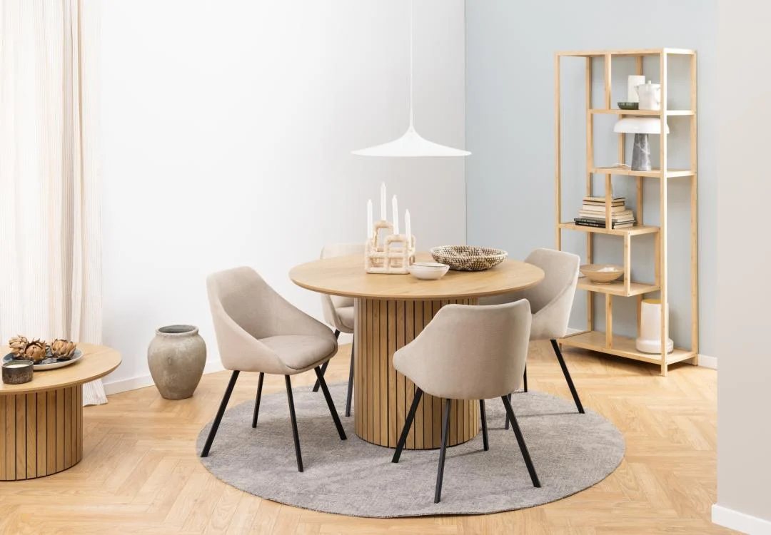Een moderne eethoek met de Eettafel Nieuwe Sfeer 120 CM Rond MDF tafel, vier beige gestoffeerde stoelen, een grijs cirkelvormig vloerkleed, open rekken met decor, een bijzettafel en zacht natuurlijk licht van een nabijgelegen raam.