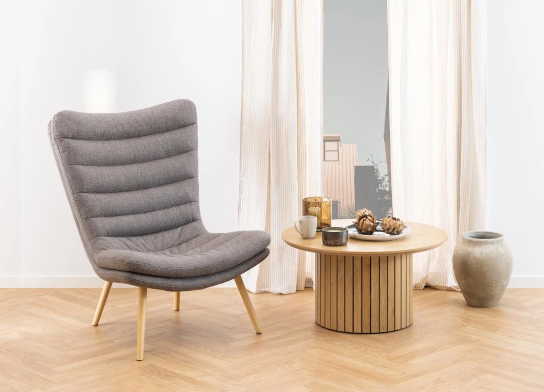 Een moderne woonkamer met een grijs gestoffeerde stoel, de Salontafel 80 CM Rond Kunststof Compact met mokken en decor, een grote keramieken vaas en hoge witte gordijnen voor een raam.