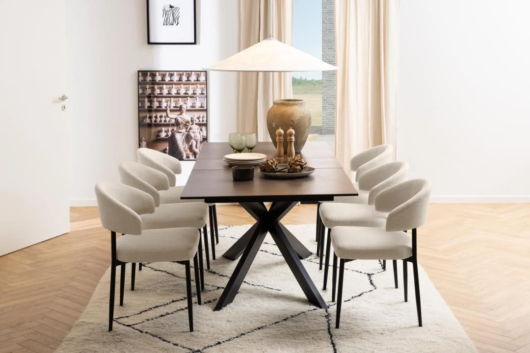 In een moderne eetkamer staat de Eettafel Nieuwe Sfeer 90 x 76 CM, een rechthoekige tafel van keramiek en staal, omringd door zes crèmekleurige stoelen. Een grote vaas, schalen en kaarsen staan in het midden; kunst siert de muur en beige gordijnen omlijsten lichte ramen.