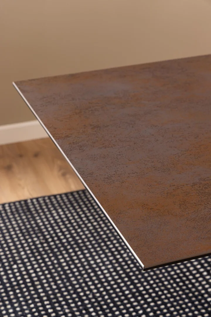 Een close-up van de hoek van de Eettafel Nieuwe Sfeer 200 x 100 CM, een dun, gestructureerd, rechthoekig tafelblad van keramiek, boven een zwart-wit geblokt tapijt en een licht houten vloer in een kamer met beige muren.