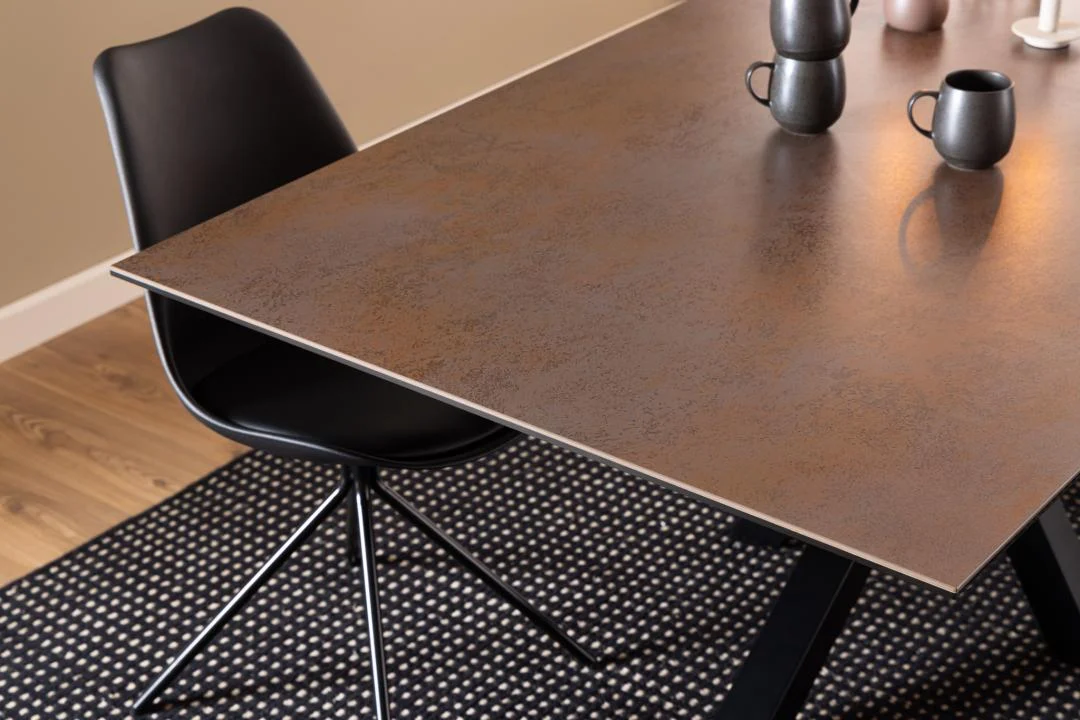 Een moderne zwarte stoel staat naast de Eettafel Nieuwe Sfeer 200 x 100 CM, een rechthoekige tafel van keramiek. Twee mokken en een vaas staan er bovenop, allemaal op een zwart-wit gedessineerd tapijt op een houten vloer.