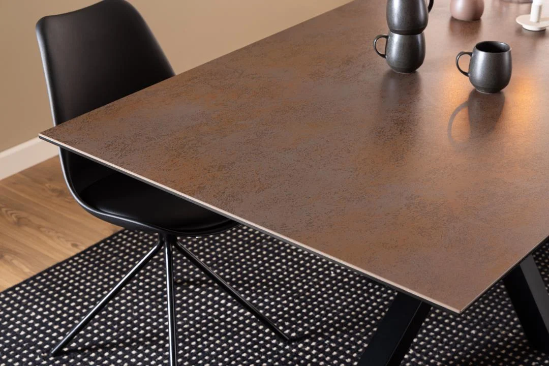 Een zwarte stoel met metalen poten staat naast de Eettafel Nieuwe Sfeer 200 x 100 CM Rechthoek Keramiek, die een bruin oppervlak met textuur heeft, drie mokken van donker keramiek, een kleine vaas en staat op een kleed met zwart en wit patroon.