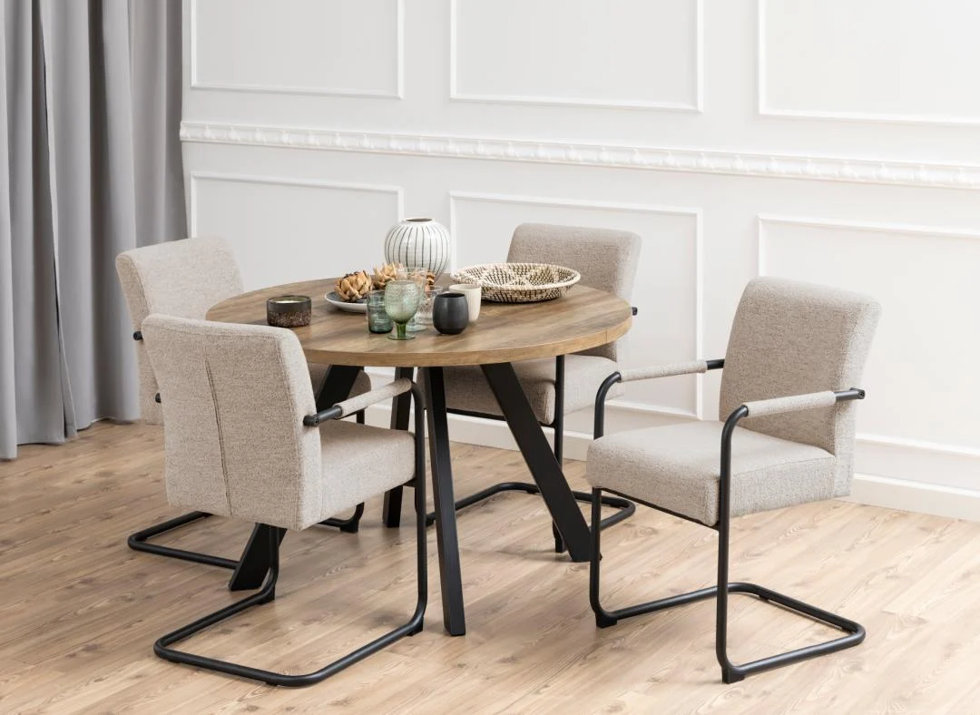 De Eettafel Zalida 120 CM Rond Eikenhout, een ronde eiken tafel met zwart stalen uitschuifbare poten, is gekoppeld aan vier beige gestoffeerde stoelen met zwarte metalen frames, op een licht houten vloer bij een wit gelambriseerde muur en grijze gordijnen. Op de tafel liggen decoraccenten.