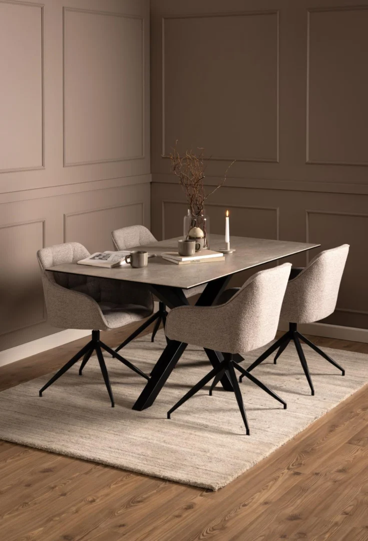 Een moderne eetkamer met de Eettafel Nieuwe Sfeer 160 x 90 CM (rechthoekig keramiek en staal), vier stoelen met kussens, een licht vloerkleed en neutraal getinte muren. Op de tafel staan een vaas met takken, een kaars, mok en boeken voor minimalistische gezelligheid.