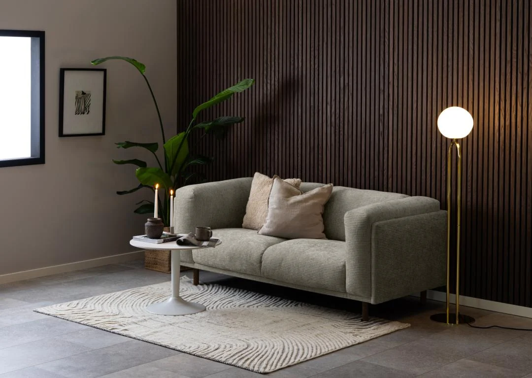 Een moderne woonkamer met een grijze bank, neutrale kussens, een Salontafel Wit 60 CM Rond Kunststof als salontafel, een vloerkleed met structuur, een hoge groene plant, een staande lamp met rond licht en een accentmuur met houten panelen.