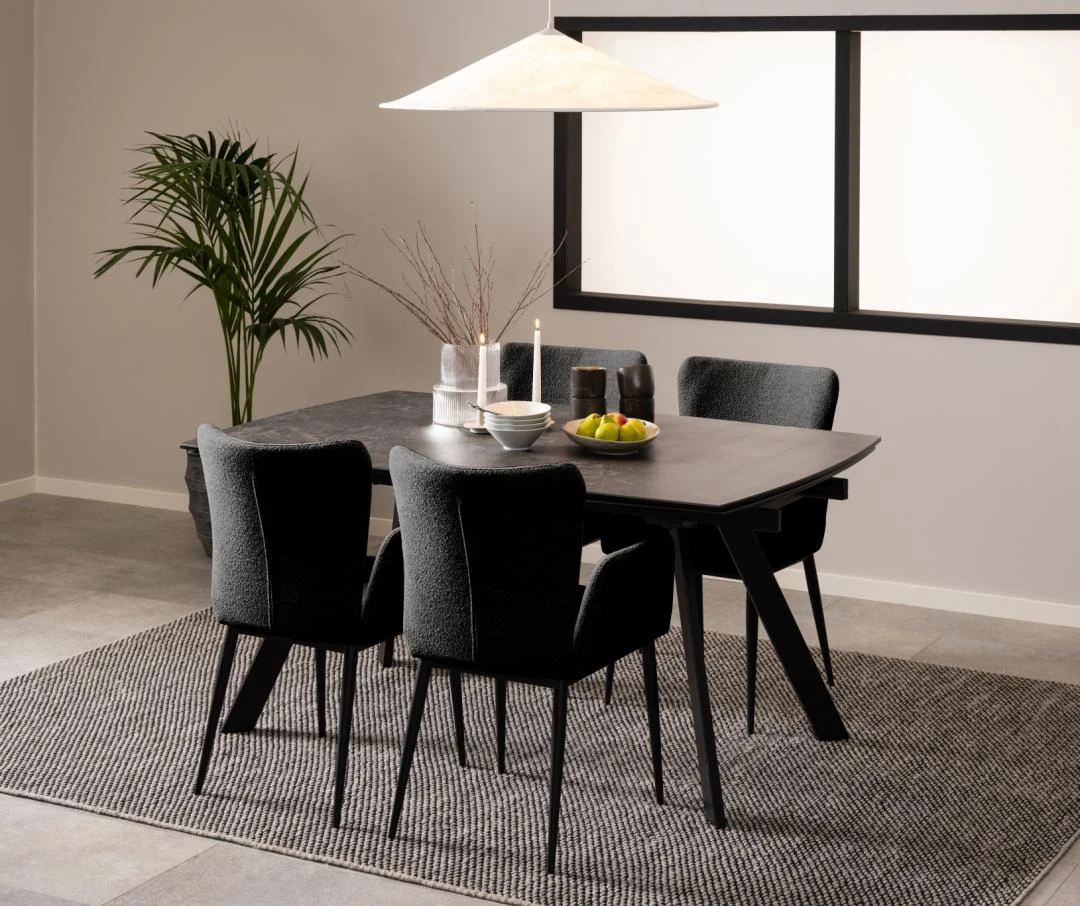 Een moderne eetkamer toont de Eettafel Nieuwe Sfeer (97 x 75 cm) met een zwart rechthoekig glazen blad, vier zwart gestoffeerde stoelen, een grijs vloerkleed met structuur, een potplant, minimalistische inrichting en een grote hanglamp in een neutraal palet.