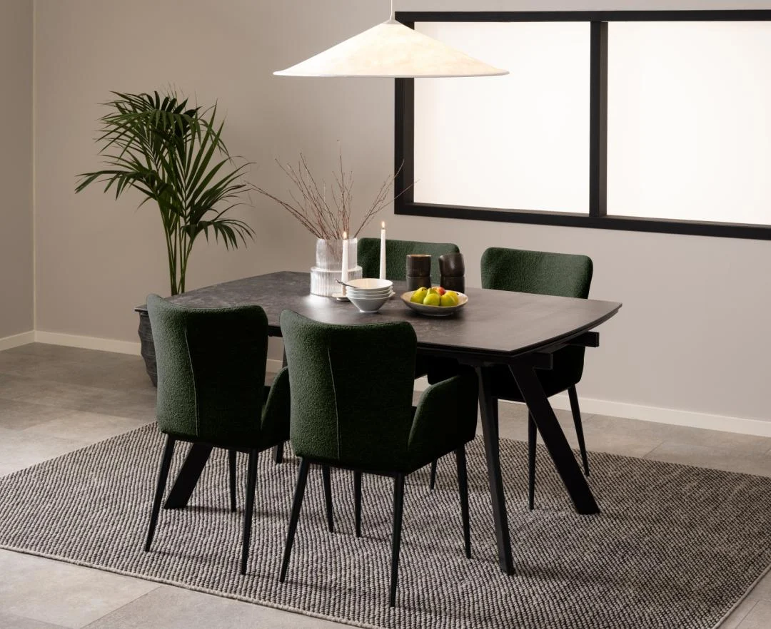 In een moderne eetkamer staat de Eettafel Nieuwe Sfeer 97 x 75 CM, een donkere rechthoekige glazen tafel, met vier groen gestoffeerde stoelen op een grijs vloerkleed. De tafel is gedecoreerd met een potplant, kaarsen en fruit, onder een grote hanglamp.